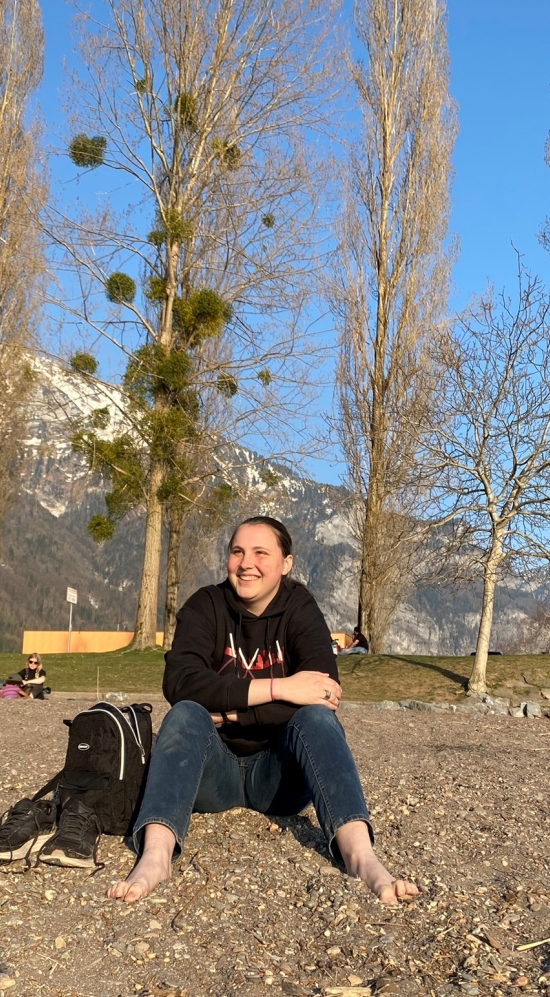 Aquila Simmons sitting with hands crossed smiling, looking away from the camera. Sitting on the ground with bald trees in the background and the Swiss Alps in a distance
