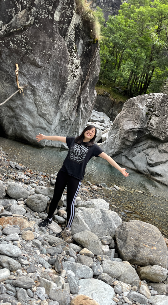 Raphi Kang poses with hands wide out in the Ticino Valley surrounded by rock formation and a forest in the background, standing on a a ground full of rocks and a stream a couple of feet away