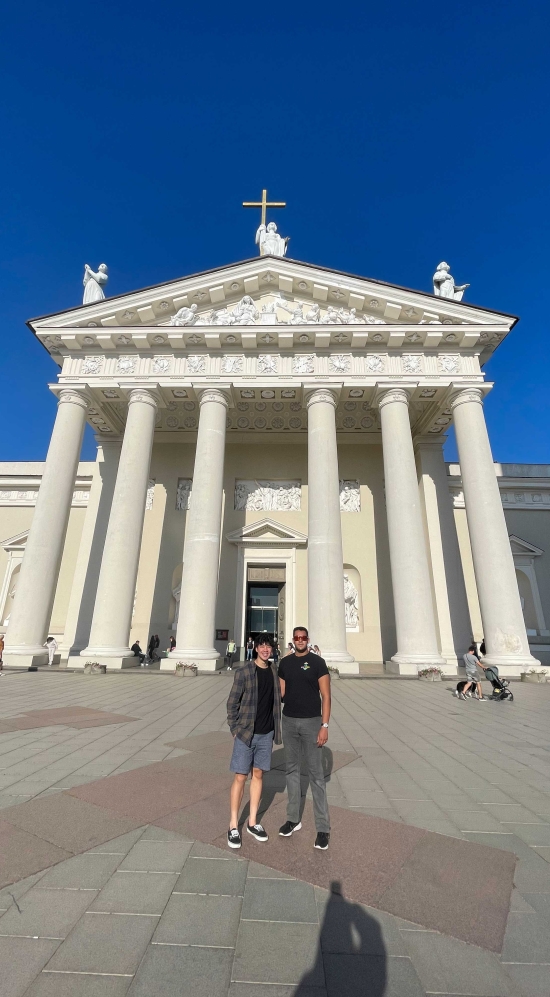 Rohan Deb and Ethan Nguyen in front of the Vilnius Cathedral thats all white and has a cross and three 3 statues atop the cathedral held by 6 large round columns