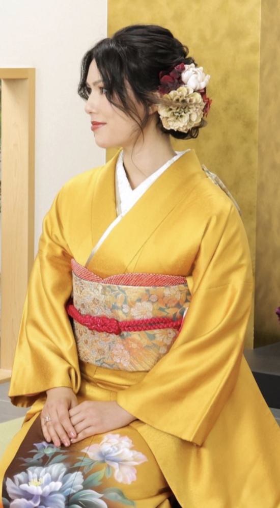 Kaili in a gold-yellow kimono in a traditional way of sitting "seiza" looking to the side to show an elaborate floral head piece that holds her hair up