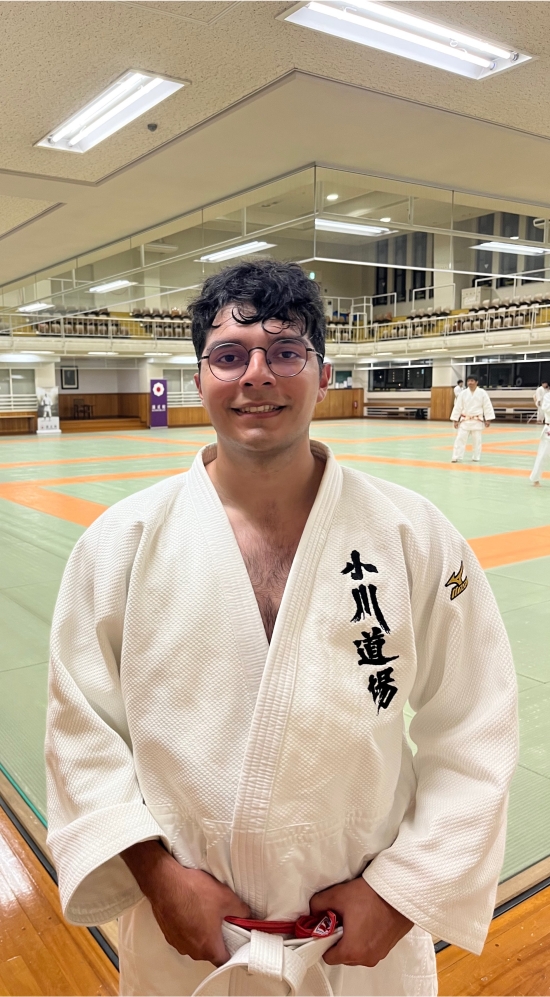 Michael Iglesias clad in a white judo uniform in a gym with a pale green mat on wooden floors with four other people in judo uniforms scattered around the judo gym on and off the pale green mat