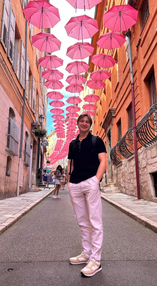 Mark Razanau posing with his hands in his pockets standing in the street in Old Town Grasse in France. The street is alley lined with opened pink umbrellas between burnt orange buildings