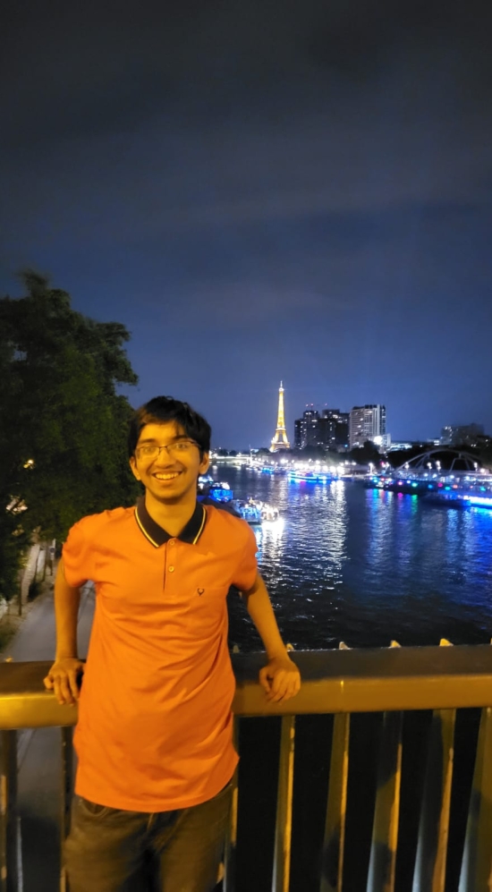 Ishan Ganguly stands on the bridge overlooking the Seine River at night with a lit Eiffel Tower in the background