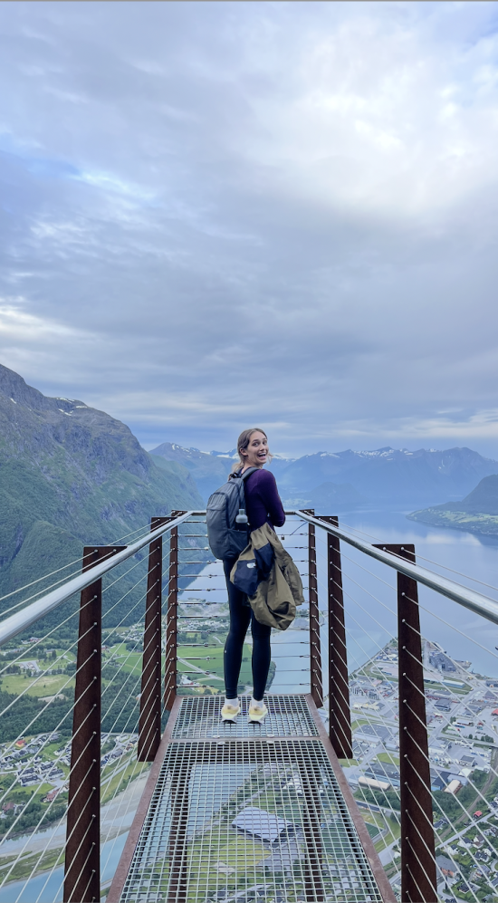 Ashley Margetts standing on a mountain viewing platform with a scenic view of mountains, water, and a town below.