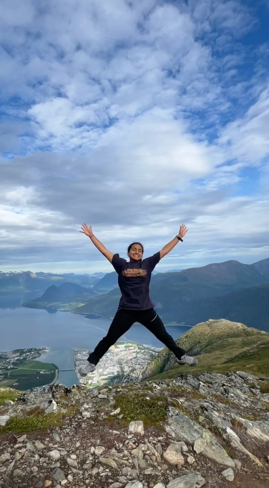 Erin Menezes in a star jump with a mountainous region in the background and a body of water between it
