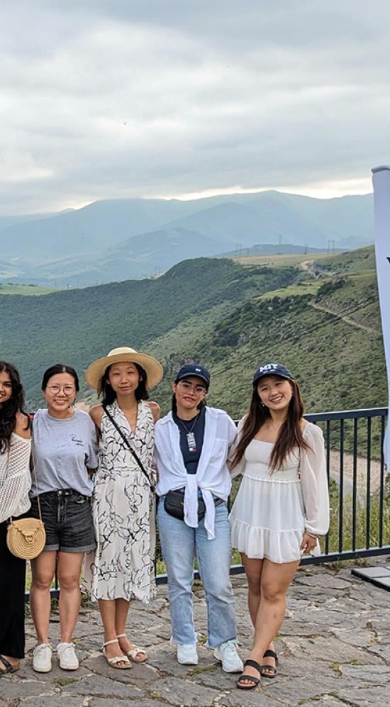 Five MIT students standing on cobbled stone groound with a railing behind them and a mountainous region in the horizon with an overcast