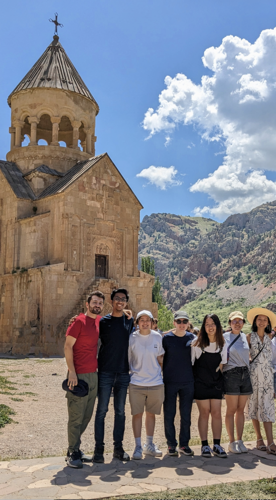 MIT students in a row in front of a monastery with a mountainous range in the back on a sunny day