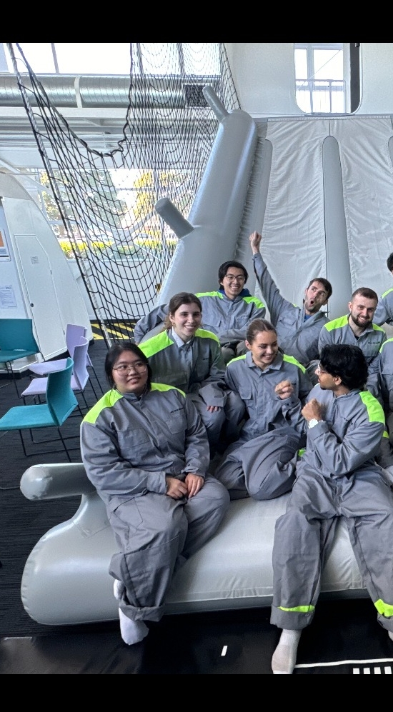 12 people in grey long sleeve onesie with lime green stripes on the shoulders seated in three rows on a grey slide indoors
