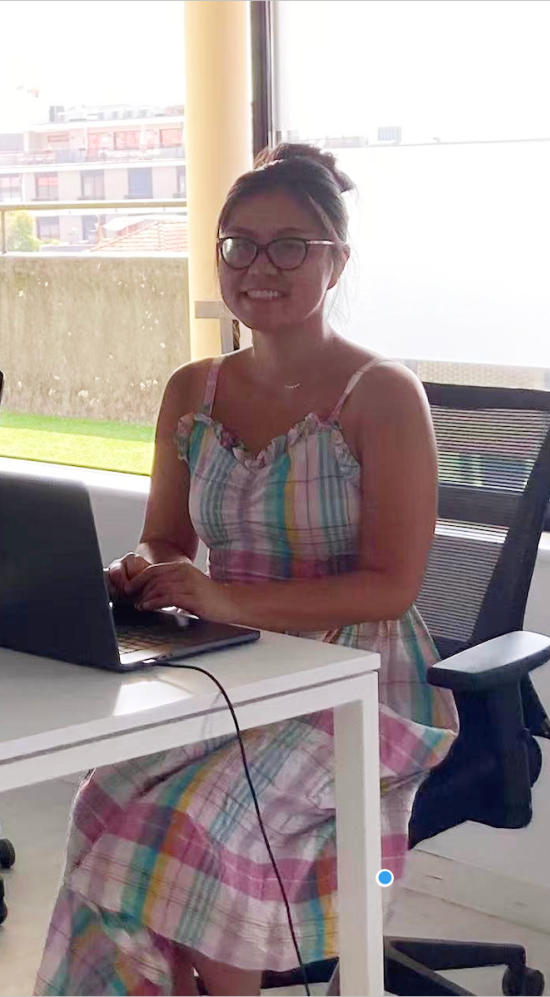 Chen Li in a plaid dress seated in front of a black laptop that's on a white table with windows behind her