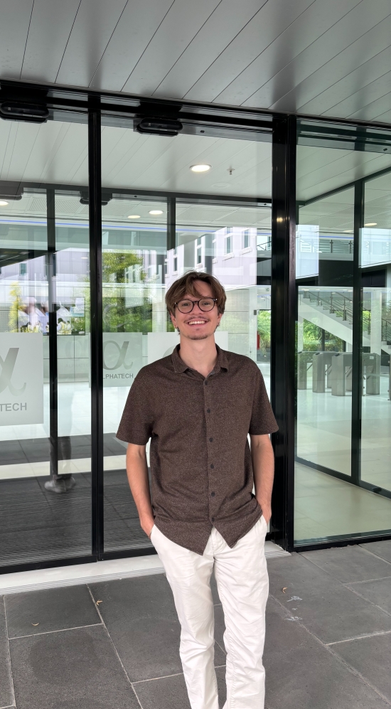 Kenny Logan with both his hands in his pockets standing outside an entrance with black framed. glass doors 