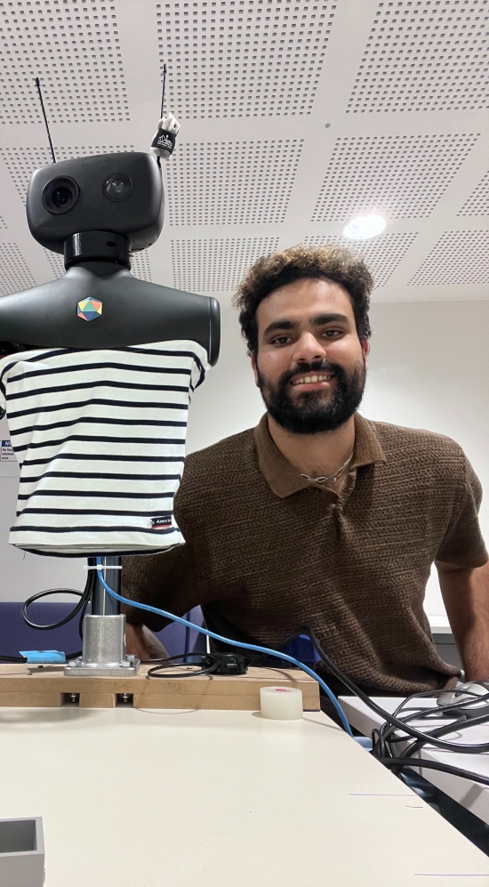Darsheep G seated on a chair with a black robot on a desk propped up wearing striped clothe as its torso