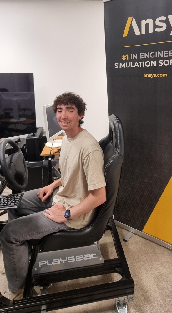 Jack seated on a chair in front of a steering wheel, keyboard and screen and to his back a roll up banner with the text "Ansys #1 Engineering Simulation Software"