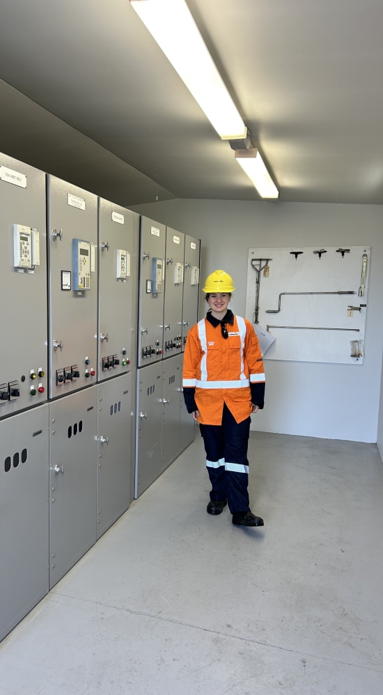 Corina A. in a orange safety suit with with yellow safety helmet next to a two-tiered grey generators