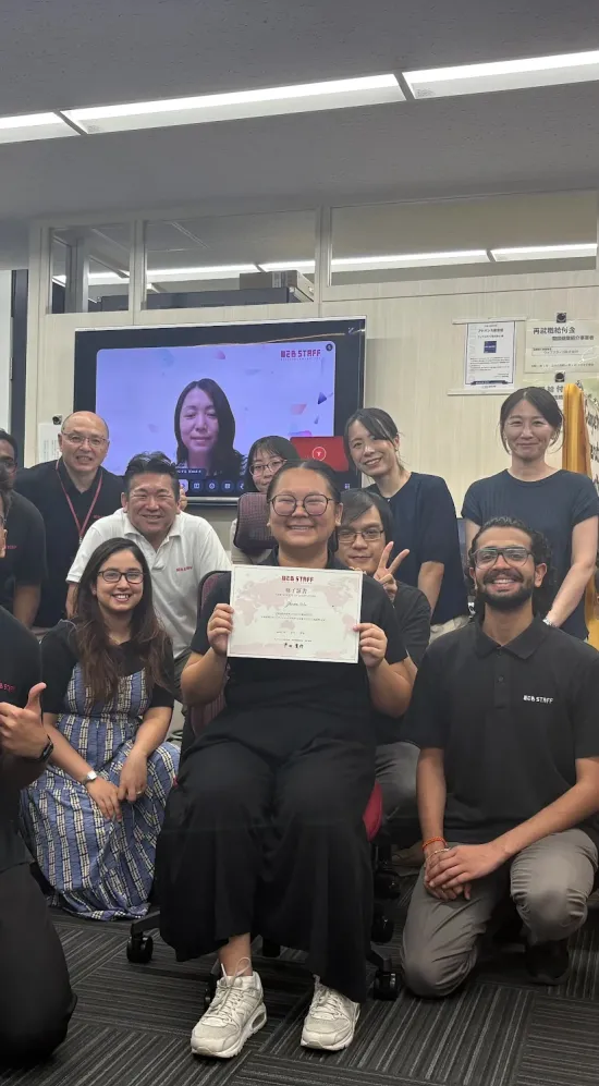 Zhiron seated and holding up a certificate surrounded by 15 colleagues in the IBJ office