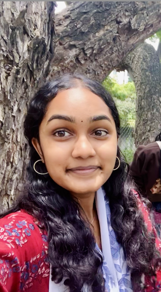 Kanna Pichappan selfie with a friend in the back who is holding up a peace sign in between two tree trunks on a sidewalk