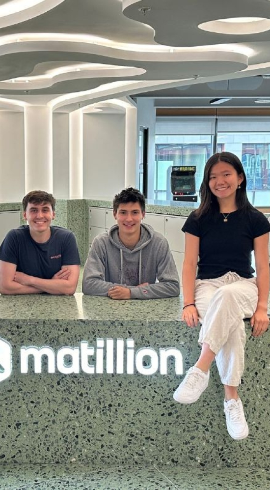 Sean Huckleberry and one other MIT student to his left seated behind a green counter with the matillion sign in the front and two MIT students seated on the counter top in between Sean and his friend.. 
