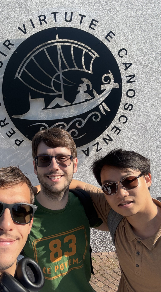 Selfie of Tri Nguyen and two others in front of a large round logo saying Per Seguir Virtute E Canoscenza