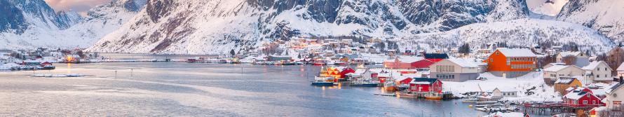 A fishing village of bright red and orange cabins, with snow-covered roofs in the city of Reine, Norway and snowy mountains in the background