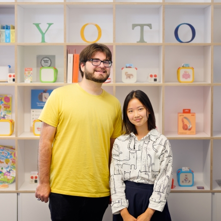 Devin Murphy and Tiffany Louie standing in front of a boxed wall filled with children's toys by Yoto in the UK