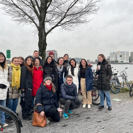 MIT Students in winter jackets standing in two rows by a river on a cloudy day in front of a barren tree with bicycles to the right and one to the left in Amsterdam