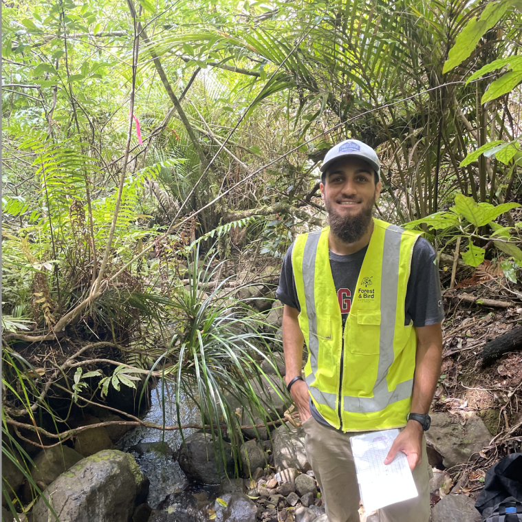 Derek Allmond in a grey cap and a yellow safety vest in a luscious green forest with a stream to the left of him as he hold one piece of paper in his hand