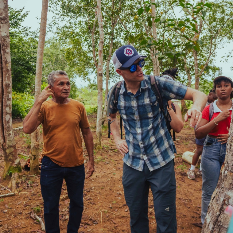 Professor Brad Olsen in a baseball cap looking at a tree, students from MIT and MUSA, surrounding him in the Amazon