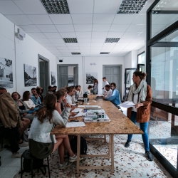 Two seated rows of MIT students sitting to the left of a long rectangle wood table while two seated at the front and another two people in the front and back standing to the right of the table in a room with floor-to-ceiling glass windows to the right with sun shining in
