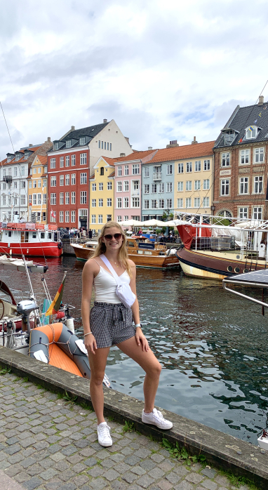 Meredith Arterburn standing next to a canal in Copenhagen, Denmark, filled with colorful boats. Across the canal there's a row of colorful four to five story buildings.