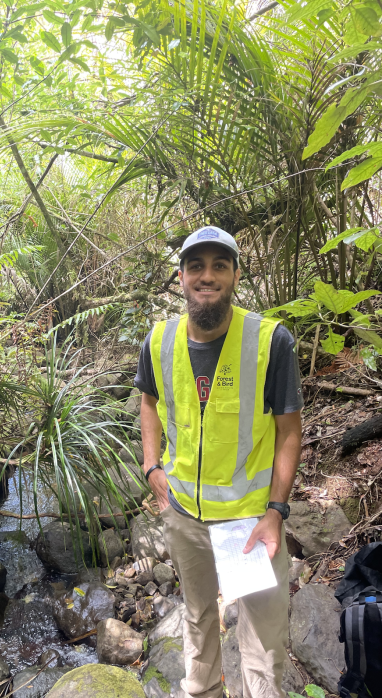 Derek Allmond in a grey cap and a yellow safety vest in a luscious green forest with a stream to the left of him as he hold one piece of paper in his hand