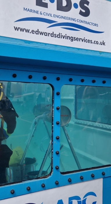 Amy Shi looking into a blue tank with someone in it wearing a wetsuit and oxygen tank under water