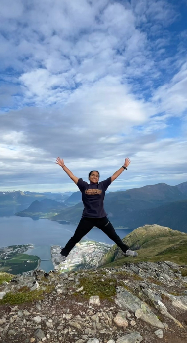 Erin Menezes in a star jump with a mountainous region in the background and a body of water between it