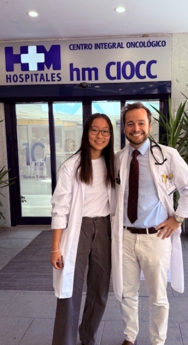 Yolanda Xie, MIT course 6, standing next to supervisor, and both wearing white lab coats in front of the entrance to HM Hospitales in Madrid, Spain