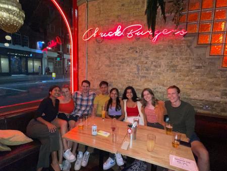 Anjali Chadha sitting in a corner table with 7 other friends at Chuck Burger Bar with a pink neon sign on a brick wall above their table that says Chuck Burgers