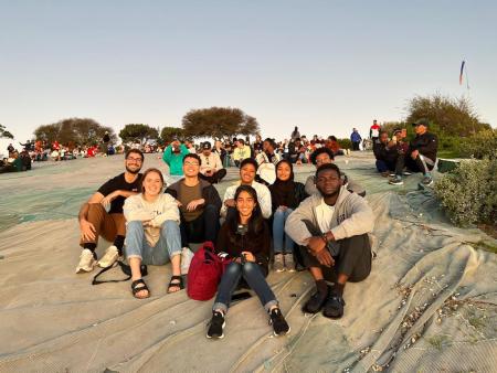 Brandon Spitzer sitting far left in a black shirt and brown pants with seven other MISTI students on a tarp on top of Signal Hill in Cape Town, South Africa during sunset with many other groups of people in the background