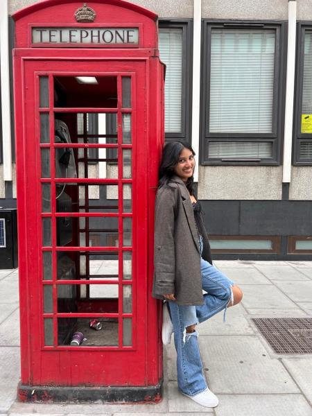 Opalina Vetrichelvan leaning on a red phone booth with one leg bent and foot against the phone booth