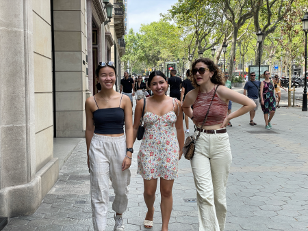 Sally Zhao walking with two friends along the central streets of Barcelona with trees lines on the sidewalk on the right and tall buildings on the left