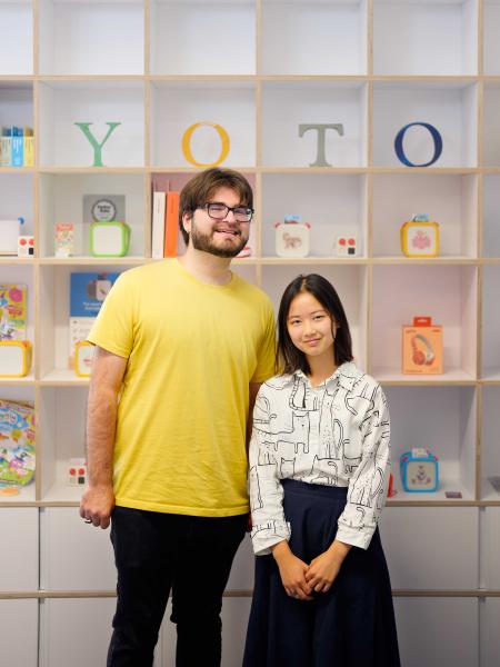 Tiffany Louie pictured with Devin Murphy in front of Yoto Limited sign, spelled using individual letters arranged in a white-cubed boxed shelf with other children toys in the grid boxes.
