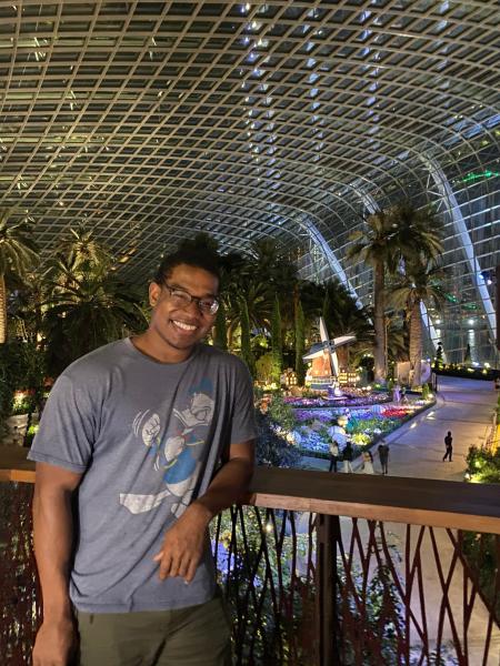 Zachary Francis leaning against a railing of the Singapore airport at night with palm trees in the background and light lit up along the path and a lit up windmill in the floor below