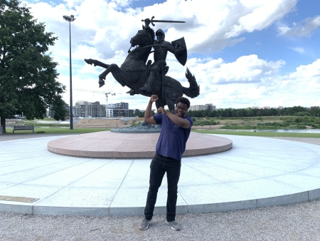 Dami Thomas in Lithuania in front of a large bronze statue with a person holding a shield and sword on a horse on two legs ready to charge