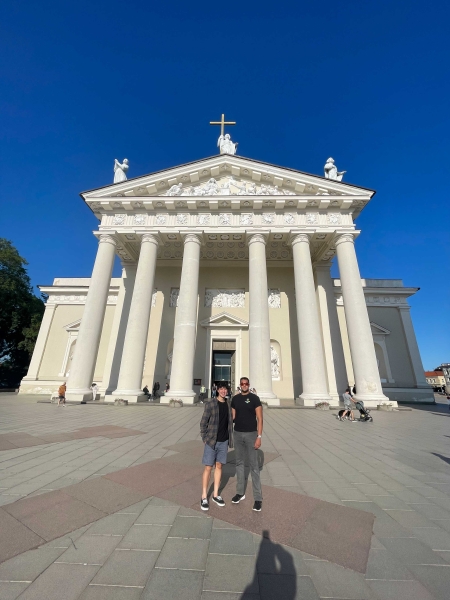 Rohan Deb and Ethan Nguyen in front of the Vilnius Cathedral thats all white and has a cross and three 3 statues atop the cathedral held by 6 large round columns