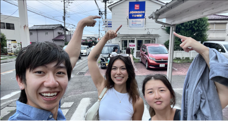 Kaili in the middle of two other MIT students in a selfie pointing to a blue sign that reads Johnson Town on a sidewalk of Tokyo