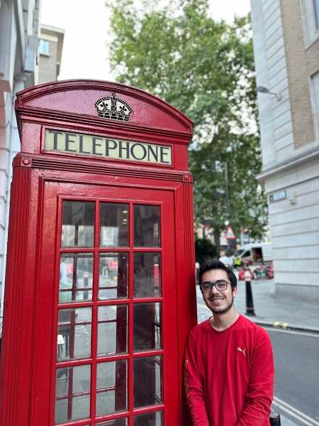 Ian Rosado wearing a red long sleeve shirt standing next to a red telephone booth in London