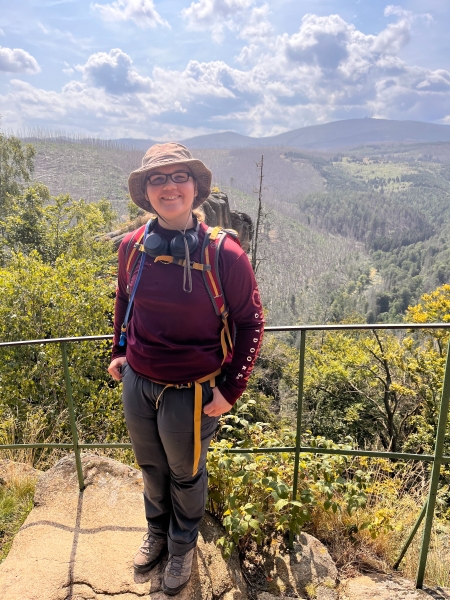 Tamara Hinderman standing on a pathwith a railing in hiking gear on a sunny day with a mountainous and green background