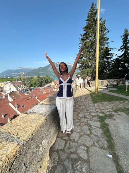 Zuri lifting both hands up on a day with clear skies, red brick roofs over the ledge to her left standing on cobbled stone path