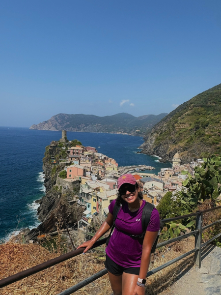 Shriya Thakur in a cap and sunglasses holding onto the railing that overlooks the sea and buildings on the coast