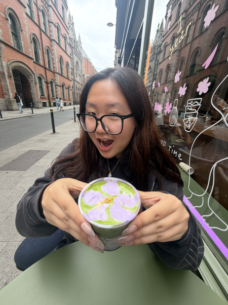 Elaine Chang holding a drink thats green with purple foam while seated at a green table on the sidewalk of London
