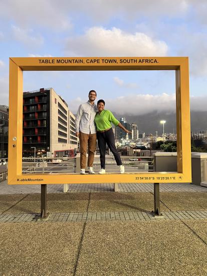 Branden Spitzer pictured behind a giant yellow frame that says Table Mountain, Cape Town, South Africa on the top of the frame with the the city scape in the background