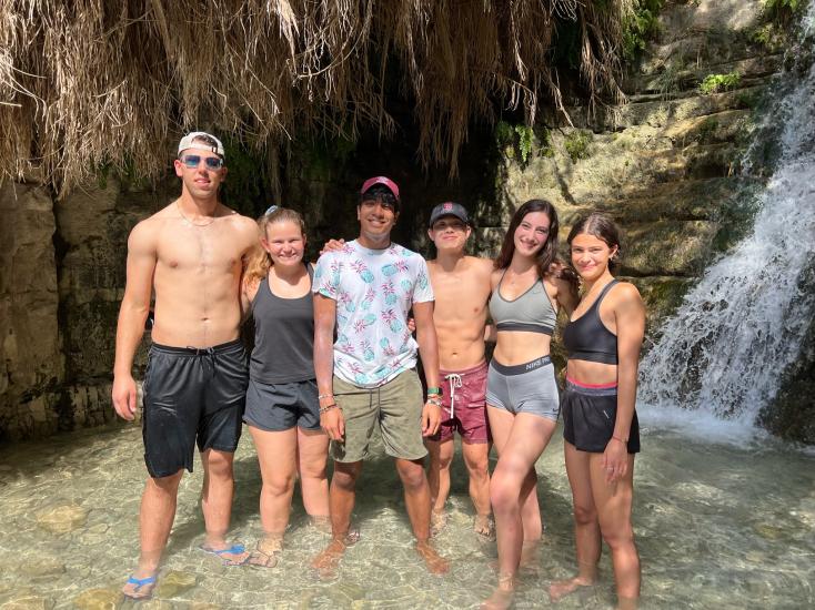 Jonathan Berger standing far left with five other MIT students standing in the shallow pool, the water reaching their ankles, of Ein Gedi Waterfall in Israel, the waterfall streaming down in the background