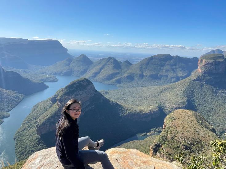 Megan Lim sitting on a rock formation with the Three Rondavel, Mpumalanga Viewpoint in South Africa. One of the three flat-rounded topped peak is seen to the right and with a birds-eye view over the Blyde River Canyon