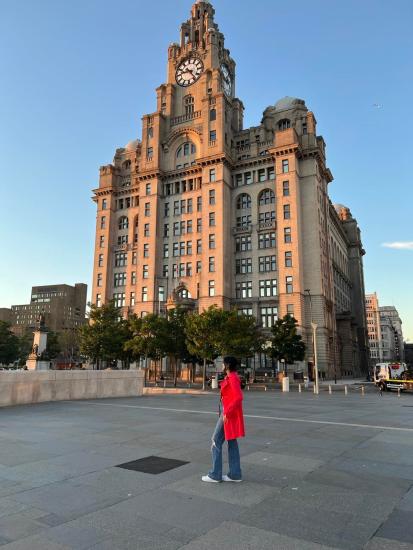 Opalina Vetrichelvan donning in a red coat and jeans, posing to her side in front of Royal Livery Building in Liverpool, UK